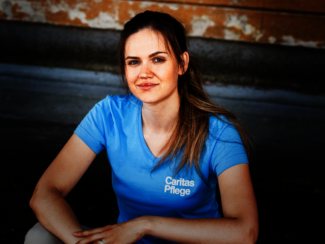 Eine junge Frau mit braunen langen Haaren und Pferdeschwanz sitzt auf einer Stiege, sie trägt ein hellblaues T-Shirt mit Caritas Logo und lächelt in die Kamera.