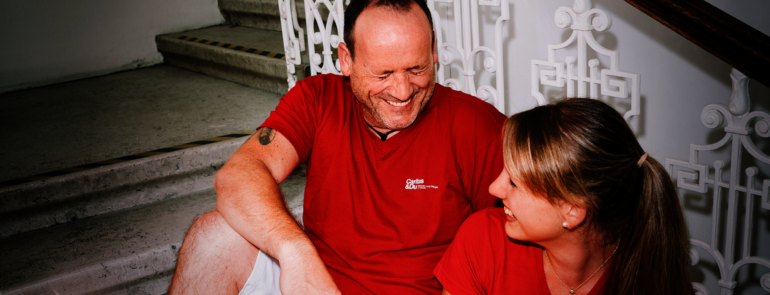 Ein Mann und eine Frau mit roten T-Shirts mit Caritas-Logo sitzen auf den Stiegen und lachen gemeinsam. Die Frau hat einen Aktenordner in der Hand.