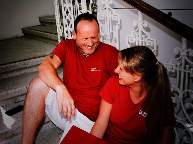 Ein Mann und eine Frau mit roten T-Shirts mit Caritas-Logo sitzen auf den Stiegen und lachen gemeinsam. Die Frau hat einen Aktenordner in der Hand.